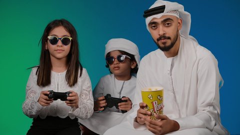 الاستمتاع بالتقنيات والتكنولوجيا الحديثة، أجواء عائلية مليئة بالسعادة والحب، ايماءات وحركات يدين تدل على التحدي باللعبة، مفهوم الانشطة الترفيهية المسلية مع الأهل، تصوير لعائلة عربية خليجية اماراتيه تقضي أوقات ممتعة في لعب الألعاب الالكترونية، خلفيه ملونه