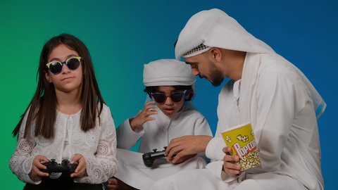 مفهوم المتعة والتسلية، أجواء عائلية مليئة بالسعادة والحب، ايماءات وحركات يدين تدل على التحدي باللعبة، مفهوم الانشطة الترفيهية المسلية مع الأهل، تصوير لعائلة عربية خليجية اماراتيه تقضي أوقات ممتعة في لعب الألعاب الالكترونية، خلفيه ملونه