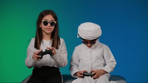 قضاء اوقات ممتعه بلعب الالعاب الالكترونيه ، مفهوم المتعة والتسلية، ايماءات وحركات يدين تدل على التحدي باللعبة، مفهوم الانشطة الترفيهية المسلية مع الأهل، تصوير لولد عربي خليجي اماراتي يحمل عصا التحكم بيده يقوم باللعب برفقة اخته، أجواء عائلية مليئة بالسعادة والحب، خلفيه ملونه