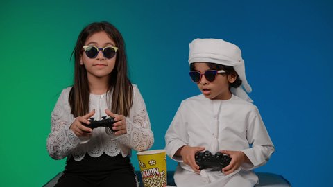 مفهوم المتعة والتسلية، ايماءات وحركات يدين تدل على التحدي باللعبة، قضاء اوقات ممتعه بلعب الالعاب الالكترونيه ، مفهوم الانشطة الترفيهية المسلية مع الأهل، تصوير لولد عربي خليجي اماراتي يحمل عصا التحكم بيده يقوم باللعب برفقة اخته، أجواء عائلية مليئة بالسعادة والحب، خلفيه ملونه