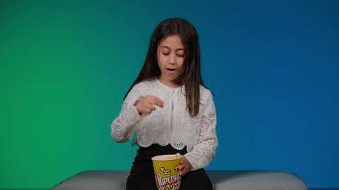 Pointing to a specific goal, having fun playing, using modern technology for entertainment, a picture of a smiling Emirati Gulf Arab girl wearing casual clothes sitting on a gray couch playing video games with expressions and gestures of integration and focus, colorful background