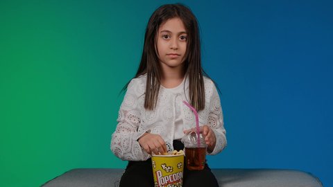Enjoying eating popcorn and drinking cold drinks, having a good time playing, using modern technology for entertainment, a picture of a smiling Emirati Gulf Arab girl wearing casual clothes sitting on a gray couch playing video games with expressions and gestures of integration and concentration, colorful background