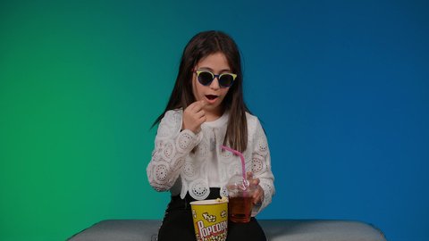 Using modern technology for entertainment, enjoying eating popcorn and drinking cold drinks, having a good time playing, a picture of a smiling Emirati Gulf Arab girl wearing casual clothes and black glasses sitting on a gray couch playing video games with expressions and gestures of integration and concentration, colorful background