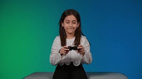 ايماءات الحماس والفوز والانتصار،استخدام التقنيات الحديثة للتسلية والترفيه، قضاء اوقات ممتعة باللعب ، تصوير لبنت عربية خليجية اماراتيه مبتسمة ترتدي الزي الكاجوال تجلس على اريكة رمادية تقوم باللعب في ألعاب الفيديو بتعابير وايماءات الاندماج والتركيز، خلفية ملونه