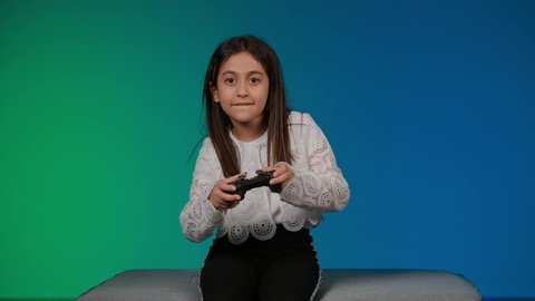 تعابير وايماءات الاندماج والتركيز،استخدام التقنيات الحديثة للتسلية والترفيه، قضاء اوقات ممتعة باللعب ، تصوير لبنت عربية خليجية اماراتيه مبتسمة ترتدي الزي الكاجوال تجلس على اريكة رمادية تقوم باللعب في ألعاب الفيديو ، خلفية ملونه