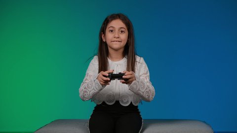 Expressions of sadness and loss, using modern technology for entertainment and amusement, having a good time playing, a photograph of a smiling Emirati Gulf Arab girl wearing casual clothes sitting on a gray couch playing video games, colorful background