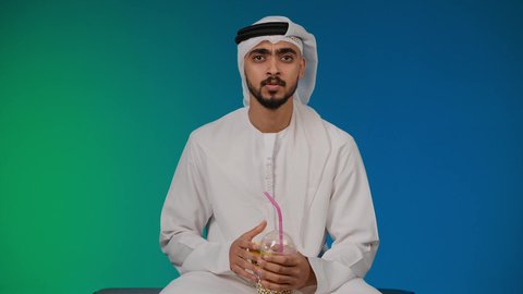 اجهزة وتقنيات الكترونية حديثة ترفيهيه, الاستمتاع بالكل البوب كورن وشرب المشروبات البارده،  قضاء وقت ممتع ومسلي، التفاعل والاندماج مع ألعاب الفيديو ، تصوير لشاب عربي خليجي اماراتي يرتدي الكندوره والغتره يجلس على كرسي مريح ويحمل بيده عصا التحكم، خلفية ملونه