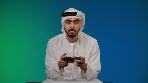 ايماءات الفوز والانتصار، قضاء وقت ممتع ومسلي، اجهزة وتقنيات الكترونية حديثة ترفيهيه, التفاعل والاندماج مع ألعاب الفيديو ، تصوير لشاب عربي خليجي اماراتي يرتدي الكندوره والغتره يجلس على كرسي مريح ويحمل بيده عصا التحكم، خلفية ملونه