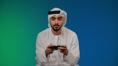 ايماءات الفشل والخساره، قضاء وقت ممتع ومسلي، اجهزة وتقنيات الكترونية حديثة ترفيهيه, التفاعل والاندماج مع ألعاب الفيديو ، تصوير لشاب عربي خليجي اماراتي يرتدي الكندوره والغتره يجلس على كرسي مريح ويحمل بيده عصا التحكم، خلفية ملونه