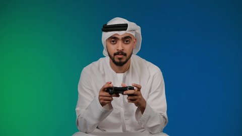 تعابير الجهل وعدم المعرفه بامر ما، قضاء وقت ممتع ومسلي، اجهزة وتقنيات الكترونية حديثة ترفيهيه, التفاعل والاندماج مع ألعاب الفيديو ، تصوير لشاب عربي خليجي اماراتي يرتدي الكندوره والغتره يجلس على كرسي مريح ويحمل بيده عصا التحكم، خلفية ملونه