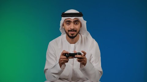 التفاعل والاندماج مع ألعاب الفيديو ، قضاء وقت ممتع ومسلي، اجهزة وتقنيات الكترونية حديثة ترفيهيه,  تصوير لشاب عربي خليجي اماراتي يرتدي الكندوره والغتره يجلس على كرسي مريح ويحمل بيده عصا التحكم، خلفية ملونه