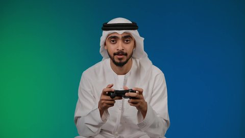 رفع قبضه اليد بايماءات الفوز ، التفاعل والاندماج مع ألعاب الفيديو ، قضاء وقت ممتع ومسلي، اجهزة وتقنيات الكترونية حديثة ترفيهيه,  تصوير لشاب عربي خليجي اماراتي يرتدي الكندوره والغتره يجلس على كرسي مريح ويحمل بيده عصا التحكم، خلفية ملونه