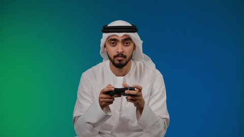 اجهزة وتقنيات الكترونية حديثة ترفيهيه, رفع اليد بايماءات الخساره ، التفاعل والاندماج مع ألعاب الفيديو ، قضاء وقت ممتع ومسلي، تصوير لشاب عربي خليجي اماراتي يرتدي الكندوره والغتره يجلس على كرسي مريح ويحمل بيده عصا التحكم، خلفية ملونه
