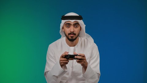التفاعل والاندماج مع ألعاب الفيديو ،اجهزة وتقنيات الكترونية حديثة ترفيهيه, ايماءات الخساره والفشل ، قضاء وقت ممتع ومسلي، تصوير لشاب عربي خليجي اماراتي يرتدي الكندوره والغتره يجلس على كرسي مريح ويحمل بيده عصا التحكم، خلفية ملونه