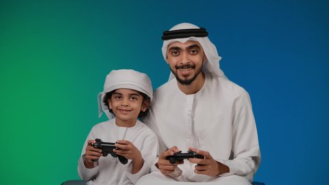 تصوير لأب وإبن إماراتي خليجي عربي يمسكان عصا التحكم و يلعبان بألعاب الفيديو، إيماءات وجه تدل على الإندماج والتفاعل مع اللعبه، المتعة و التسلية بألعاب الفيديو، ألعاب البلايستيشن الترفيهية، اللعب مع العائلة , خلفية ملونه