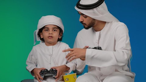 المتعة و التسلية بألعاب الفيديو،إيماءات وجه تدل على الإندماج والتفاعل مع اللعبه، تصوير لأب وإبن إماراتي خليجي عربي يمسكان عصا التحكم و يلعبان بألعاب الفيديو، ألعاب البلايستيشن الترفيهية، اللعب مع العائلة , خلفية ملونه