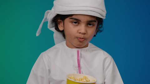 شرب المشروبات البارده، برامج الكمبيوتر الترفيهية، اكل البوب كورن، قضاء وقت ممتع بلعب ألعاب الفيديو، الاستمتاع بالتقنيات والتكنولوجيا الحديثة، مفهوم المتعة والتسلية، تصوير مقرب لطفل عربي خليجي اماراتى مبتسم  يمسك بيده عصا اللعب، خلفية ملونه