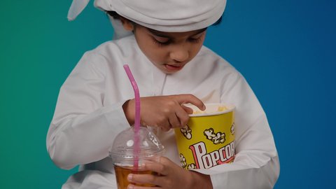 الاستمتاع بالتقنيات والتكنولوجيا الحديثة، مفهوم المتعة والتسلية،شرب المشروبات البارده، برامج الكمبيوتر الترفيهية، اكل البوب كورن، قضاء وقت ممتع بلعب ألعاب الفيديو، تصوير مقرب لطفل عربي خليجي اماراتى مبتسم  يمسك بيده عصا اللعب، خلفية ملونه