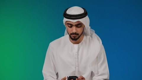 Taking commemorative selfies via mobile, making a video call via social media applications and websites, using modern devices and technologies, photographing a young Emirati Gulf Arab man wearing a kandura and ghutra holding a mobile phone in his hand, colorful background