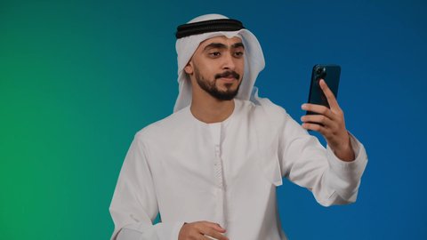 اجراء مكالمة فيديو عبر تطبيقات ومواقع السوشال ميديا، التقاط صور سيلفي تذكارية عبر الجوال،استخدام الأجهزة والتقنيات الحديثة، تصوير لشاب عربي خليجي اماراتي يرتدي الكندوره والغتره يمسك بيده الجوال ، خلفية ملونه