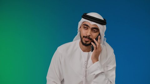 Setting a specific goal, making a video call via social media applications and websites, taking commemorative selfies via mobile, using modern devices and technologies, photographing a young Emirati Gulf Arab man wearing a kandura and ghutra holding a mobile phone in his hand, a colorful background
