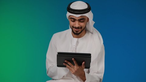 تصوير مقرب لرجل عربي خليجي إماراتي يرتدي الغترة والكندورة يقوم باستخدام الجهاز اللوحي المحمول بإيماءات الفرح والسرور، دمج الأجهزة التقنية الحديثة والتكنولوجيا بالحياة اليومية ، الدردشة والتواصل مع الأهل و الأصدقاء،تصفح مواقع التواصل الاجتماعي، خلفيه ملونه