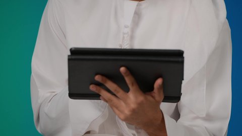 دمج الأجهزة التقنية الحديثة والتكنولوجيا بالحياة اليومية ، الدردشة والتواصل مع الأهل و الأصدقاء،تصفح مواقع التواصل الاجتماعي، تصوير مقرب لرجل عربي خليجي إماراتي يرتدي الغترة والكندورة يقوم باستخدام الجهاز اللوحي المحمول بإيماءات الفرح والسرور،  خلفيه ملونه