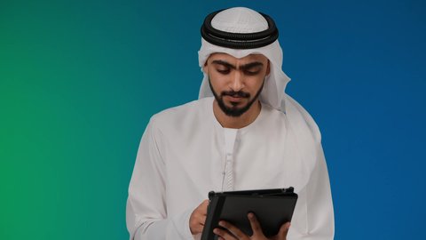 الدردشة والتواصل مع الأهل و الأصدقاء،تصفح مواقع التواصل الاجتماعي، تصوير مقرب لرجل عربي خليجي إماراتي يرتدي الغترة والكندورة يقوم باستخدام الجهاز اللوحي المحمول بإيماءات الفرح والسرور، دمج الأجهزة التقنية الحديثة والتكنولوجيا بالحياة اليومية ، خلفيه ملونه