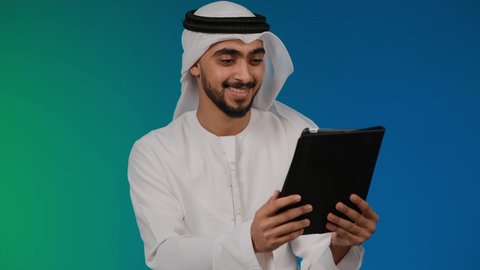 تصفح مواقع التواصل الاجتماعي، الدردشة والتواصل مع الأهل و الأصدقاء، تصوير مقرب لرجل عربي خليجي إماراتي يرتدي الغترة والكندورة يقوم باستخدام الجهاز اللوحي المحمول بإيماءات الفرح والسرور، دمج الأجهزة التقنية الحديثة والتكنولوجيا بالحياة اليومية ، خلفيه ملونه