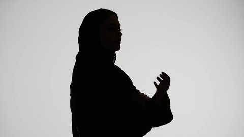 The concept of elegance and attention to one's appearance, modern, elegant veiled fashion, silhouette of an Emirati Gulf Arab woman wearing an abaya and hijab, standing upright with her arms crossed, white background.