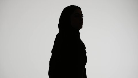 Modern, elegant, and sophisticated hijab fashion. The concept of elegance and attention to one's outward appearance. A silhouette of an Emirati Gulf Arab woman wearing an abaya and hijab, standing upright with her arms crossed, against a white background.