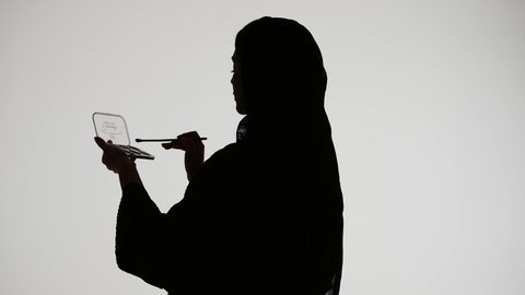 إستخدام إحدى أدوات التجميل ( فرشاة ظل العيون ) ، التسويق لمنتجات التجميل ، تصوير ظلي لمرأة عربية خليجية اماراتيه ترتدي العباءه والحجاب ، الإهتمام بالجمال والمظهر الخارجي ، أدوات مكياج تتوريال ، خلفية بيضاء