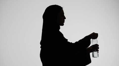 أهمية شرب الماء النقي للمحافظة على بشرة نضرة، المحافظة على سلامة الجهاز المناعي، التخلص من سموم الجسم، تصوير ظلي لامراه عربي خليجيه اماراتيه ترتدي العباء والحجاب ،تقوم بشرب المياه من عبوة بلاستيكية، خلفية بيضاء