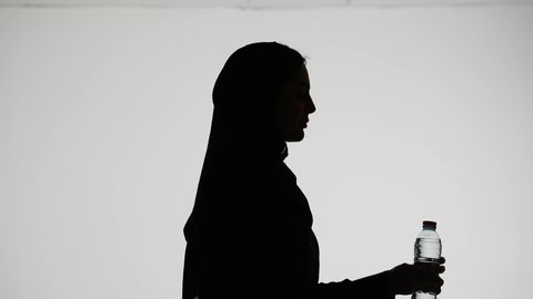 Detoxifying the body, maintaining a healthy immune system, the importance of drinking pure water for healthy skin, silhouette of an Emirati Gulf Arab woman wearing an abaya and hijab, drinking water from a plastic bottle, white background