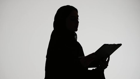 مفهوم التعليم والثقافة ،  متابعة الخطط والمشاريع ، قراءة الملاحظات والمعلومات ، كتابة الأبحاث والتقارير ، تصوير ظلي لمرأة عربية خليجية اماراتيه ترتدي العباءه والحجاب ، تمسك بيدها أوراق خاصة بالعمل ، خلفية بيضاء