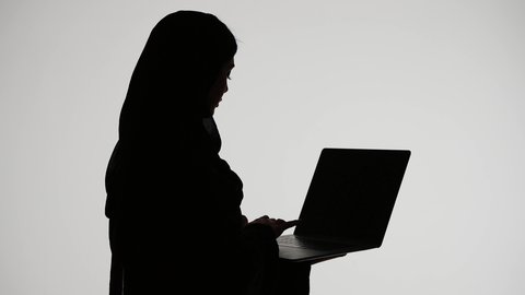 دمج التكنولوجيا الحديثه فى الحياه اليوميه ، اجراء مكالمه فيديو ،انجاز مهام العمل بإستخدام الحاسوب المحمول،إستخدام اجهزة تقنية حديثة ،وسائل التواصل الإجتماعي ، تصوير ظلي لمرأة عربية خليجية اماراتيه ترتدي العباءه والحجاب تمسك بيدها جهاز اللابتوب ، خلفية بيضاء