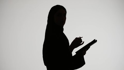انجاز مهام العمل بإستخدام التابلت ،إستخدام اجهزة تقنية حديثة ،دمج التكنولوجيا الحديثه فى الحياه اليوميه ، وسائل التواصل الإجتماعي ، اجراء مكالمه هاتفيه ،تصوير ظلي لمرأة عربية خليجية اماراتيه ترتدي العباءه والحجاب تمسك بيدها جهاز التابلت والقلم ، خلفية بيضاء