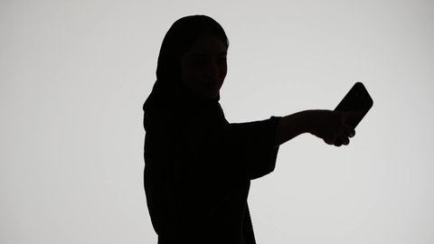 Silhouette of an Emirati Gulf Arab woman wearing an abaya and hijab, holding a mobile phone, taking a selfie, social media, using modern technology, conducting a live broadcast, making a phone call, white background