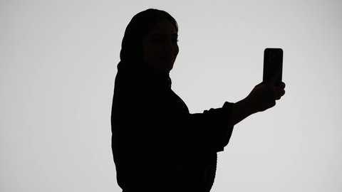 Taking commemorative selfies using a mobile phone, social media, using modern technology, conducting a live broadcast, making a phone call, a silhouette of an Emirati Gulf Arab woman wearing an abaya and hijab, white background