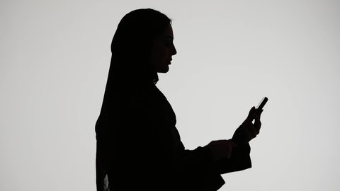 إستخدام اجهزة تقنية حديثة ،  اجراء بث مباشر ،التقاط صور سيلفي تذكاريه بإستخدام الهاتف المحمول ، اجراء مكالمه هاتفيه ،تصوير ظلي لمرأة عربية خليجية اماراتيه ترتدي العباءه والحجاب ،وسائل التواصل الإجتماعي ، خلفية بيضاء