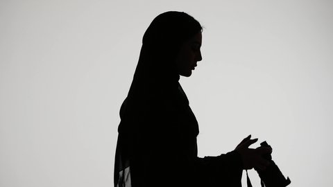 اتقان مهنه التصوير ، مصوره اماراتيه محترفه ، إلتقاط الصور التذكارية ، تصوير ظلي لامراه عربيه خليجيه اماراتيه ترتدي العباءه والحجاب، تحمل بيدها كاميرا تصوير عالية الدقة ، خلفية بيضاء