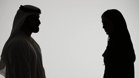 A quarrel between spouses, a crisis in family relationships, family problems and domestic conflict, a woman defending herself, an Emirati man wearing a ghutra and kandura hitting and abusing his wife in the face, a silhouette of an Arab Gulf Emirati couple arguing with each other with angry gestures, white background