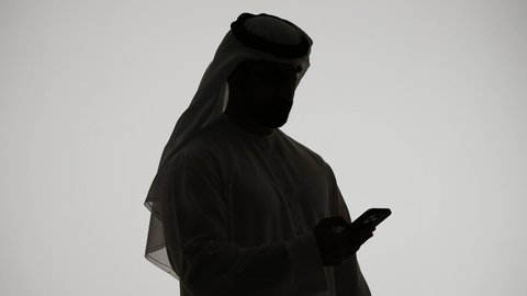 Integrating modern technologies into daily life, online chatting, making a video call using a mobile phone, managing business and achieving goals online, completing tasks remotely, silhouette of an Emirati Gulf Arab man holding a mobile phone, white background