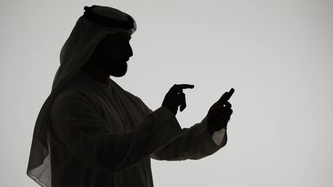 إجراء مكالمة فيديو بإستخدام الهاتف المحمول ، ادارة الاعمال وتحقيق الأهداف أونلاين، الدردشه عبر الانترنت، دمج التقنيات الحديثه فى الحياه اليوميه ، إنجاز المهام عن بعد، تصوير ظلي لرجل عربي خليجي اماراتي يمسك بيده الجوال ، خلفية بيضاء
