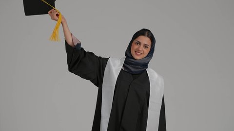 مفهوم الاجتهاد والتفوق، إنهاء الدراسة الجامعية، إيماءات الفرح والسرور، الشعور بالفخر والثقه بالنفس ، تحقيق حلم النجاح, الاحتفال بالتخرج من الجامعة، بورترية لطالبة عربية خليجية اماراتيه محجبة ترتدي روب التخرج ترفع قبعه التخرج بايديها، خلفية رمادية