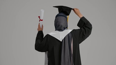 الشعور بالفخر والثقه بالنفس ، تحقيق حلم النجاح, إنهاء الدراسة الجامعية، مفهوم الاجتهاد والتفوق ، إيماءات الفرح والسرور، الاحتفال بالتخرج من الجامعة، بورترية من الخلف لطالبة عربية خليجية اماراتيه محجبة ترتدي روب التخرج ، ترفع وثيقه النجاح وقبعه التخرج بايديها، خلفية رمادية
