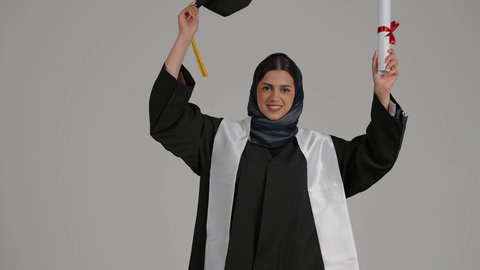 Completing university studies, the concept of diligence and excellence, gestures of joy and happiness, feeling proud and self-confident, achieving the dream of success, celebrating university graduation, close-up portrait of a veiled Emirati Gulf Arab student wearing a graduation robe, raising the success document and graduation cap in her hands, gray background