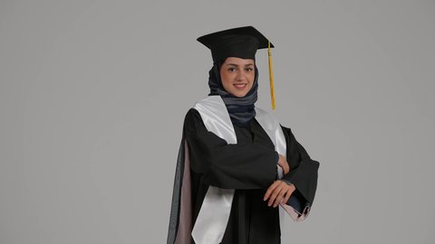 تحقيق حلم النجاح، الاحتفال بالتخرج من الجامعة، الشعور بالفخر والثقه بالنفس ، إنهاء الدراسة الجامعية، مفهوم الاجتهاد والتفوق ، بورترية لطالبة عربية خليجية اماراتيه محجبة ترتدي روب التخرج والقبعة تقف بايدي متقاطعه، خلفية رمادية
