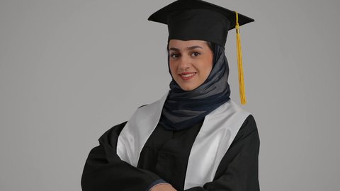 لشعور بالفخر والثقه بالنفس ، إنهاء الدراسة الجامعية، تحقيق حلم النجاح, الاحتفال بالتخرج من الجامعة، مفهوم الاجتهاد والتفوق ، بورترية مقرب لطالبة عربية خليجية اماراتيه محجبة ترتدي روب التخرج والقبعة تقف بايدي متقاطعه، خلفية رمادية