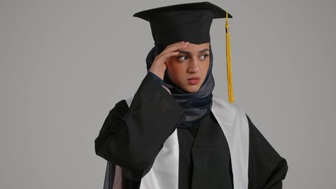الحاجه للعلاج وطلب المساعده ، إنهاء الدراسة الجامعية، تحقيق حلم النجاح, الاحتفال بالتخرج من الجامعة، مفهوم الاجتهاد والتفوق ، بورتريةمقرب لطالبة عربية خليجية اماراتيه محجبة ترتدي روب التخرج والقبعة تضع يدها على رأسها بإيماءات الدوار و الألم، خلفية رمادية